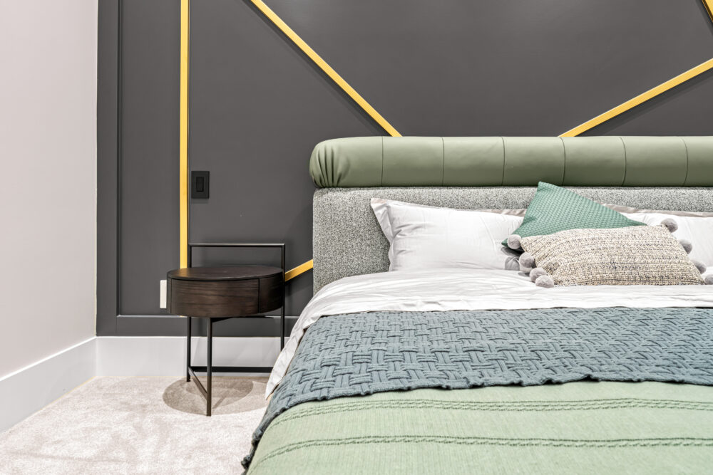 Stylish modern bedroom featuring a queen-size bed with a green velvet headboard, patterned throw pillows, and a textured grey blanket, set against a grey wall with geometric yellow accents
