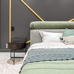 Stylish modern bedroom featuring a queen-size bed with a green velvet headboard, patterned throw pillows, and a textured grey blanket, set against a grey wall with geometric yellow accents
